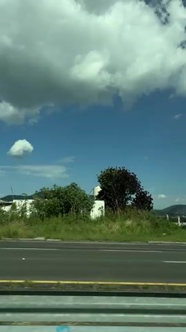 The video captures a roadside view from a moving vehicle in Santa Cruz Tehuixpango, Mexico. The scene is dominated by a bright blue sky filled with fluffy white and dark gray clouds, suggesting a partly cloudy day. The time appears to be daytime, likely late morning or early afternoon given the brightness.

The foreground shows a paved road with white lane markings and a yellow stripe, indicating a highway or major road. A metal guardrail is visible along the bottom edge of the frame. Beyond the road, a grassy embankment is seen, with some scattered white posts and fencing. In the middle ground, rolling green hills and mountains form the backdrop, with some signs of civilization like buildings and trees interspersed. A large billboard advertising "JEEP" is prominently displayed on the right side of the frame, featuring an image of a Jeep vehicle. A silver SUV is seen driving on the highway in the same direction as the viewer.

Further in the distance, there are indications of construction with a concrete structure featuring multiple pillars. The overall impression is a sunny day with dramatic cloud formations, typical of a rural or semi-rural setting in Mexico. The presence of the highway and the billboard suggests a location with some commercial activity, likely along a travel route. There are no people clearly visible in the frame. The emotions conveyed are neutral and observational, focusing on the landscape and the journey.