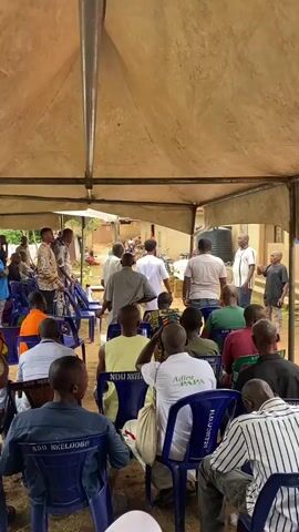 The video shows a gathering of people, mostly men, sitting on blue chairs under a beige tent. Some men are standing and appear to be speaking to the seated individuals. One man in a colorful patterned shirt is standing and gesturing. In the background, there are buildings and trees. A white coffin with gold trim is visible on the right side of the frame, resting on bricks. Several people are looking towards the coffin. Some of the seated individuals are wearing white shirts with text on them, including "Adieu PAPA". The overall atmosphere appears to be somber.