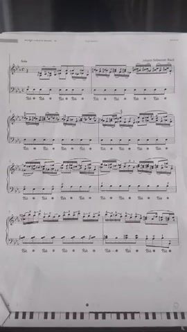This video shows a close-up, slightly angled shot of a piece of sheet music by Johann Sebastian Bach titled "Aria." The music is laid out on a dark surface, possibly a piano or a music stand, with a portion of a keyboard visible at the bottom of the frame. The lighting is somewhat dim, causing some of the details on the music sheet to be slightly obscured. The camera remains static throughout the short clip, focusing on the musical score. There are no people or other significant objects visible in the frame besides the sheet music and the keyboard.