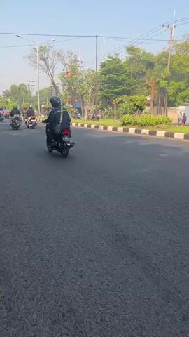 The video shows a paved road with several motorcycles and riders. The road appears to be asphalt with a white and black striped curb. There are trees and utility poles visible along the side of the road. One rider in the foreground is wearing a black helmet and a black jacket and is on a black motorcycle. Other motorcycles are also visible in the background. The sky is clear and the lighting suggests it is daytime, likely morning or late afternoon due to the sun angle. The scene appears to be in an urban or suburban area.