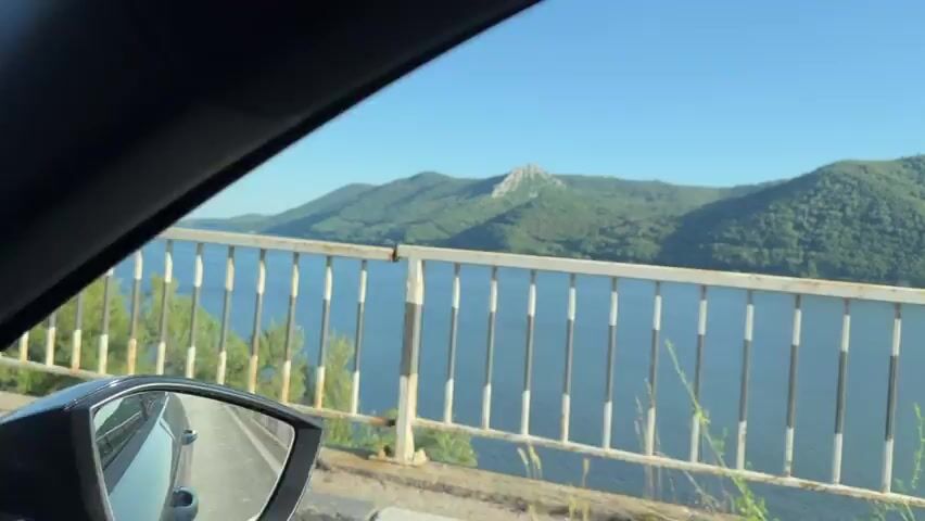 The video displays a moving shot from a vehicle, initially showing a metal railing with black and white striped posts in the foreground. Beyond the railing, a wide body of water extends, with green, tree-covered mountains visible in the distance under a clear blue sky. One mountain features a prominent, lighter-colored, rocky peak. As the camera continues to move, the railing gives way to a low stone wall, with green foliage appearing between the wall and the water. The view of the water and distant mountains remains consistent throughout the clip.