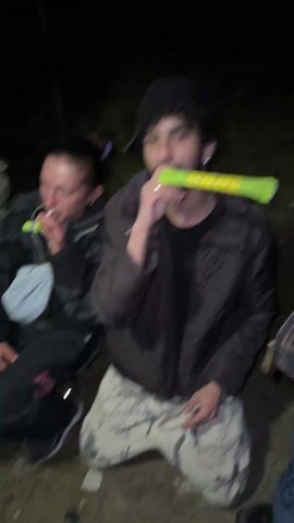 The video shows three people outdoors at night. A person with dark hair and a beard, wearing a black beanie, brown jacket, and camouflage pants, is blowing a party horn. To the left of this person is a woman with her hair pulled back, wearing a grey patterned jacket and a small light-colored bag across her chest. She also holds a party horn. To the right of the person in the brown jacket is another person partially visible, wearing a jacket and blue jeans. The setting appears to be a casual outdoor gathering. The background is dark and indistinct, possibly showing some vegetation. A second party horn is flying through the air above the central person.
