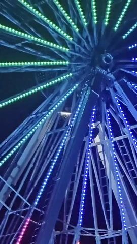 This is a close-up, low-angle, vertical shot of a Ferris wheel at night. The Ferris wheel is illuminated with rows of colored LED lights that change from green to blue to purple. The lights are arranged in radial lines extending from the center of the wheel. The structure of the Ferris wheel is visible, including metal beams and a ladder. The sky is dark and appears to be black. The video shows the lights cycling through their color sequence.