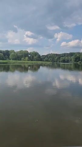 A body of water is visible in the foreground and middle ground. The water is calm, with reflections of the sky and trees. The sky is partly cloudy, with white and gray clouds against a blue sky. Trees line the far bank of the water, forming a dense green wall. The overall impression is one of natural beauty and tranquility.