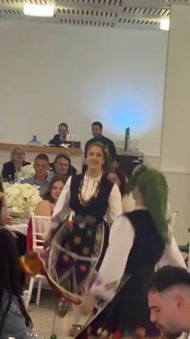 The video captures a scene within what seems to be a wedding reception or a similar formal gathering, where traditional Balkan folk dancers are performing amidst seated guests. Two women, clad in colorful folk costumes with green head coverings, are seen dancing. They are soon joined by two men, also in traditional attire, including black vests and hats. The dancers move around tables decorated with white flower arrangements, while the guests, seated in white chairs, observe the performance. The environment is a brightly lit hall with a neutral color scheme, suggesting a modern indoor event space. The camera angle is positioned among the guests, providing a close view of the dancers and audience interaction.