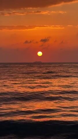 The video shows a sunset over the ocean. The sun is a bright orange orb near the horizon, casting a warm glow across the sky and water. There are some clouds in the sky, tinged with orange and pink. A large ship is visible on the horizon, silhouetted against the setting sun. The ocean waves are gently rolling towards the shore. The overall scene is peaceful and serene.