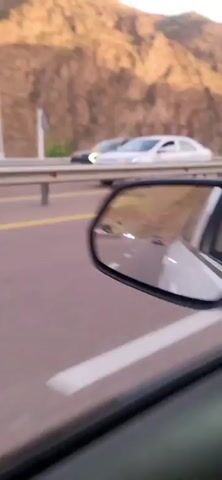 The video shows a perspective from inside a moving vehicle, likely a car. The view is of a road with a yellow line separating lanes, and a guardrail runs alongside. Beyond the guardrail, there is vegetation and a steep, rocky mountain face. The sun is casting shadows on the mountain, indicating it is either morning or late afternoon. A silver sedan passes by in the opposite direction, revealing a driver inside. The video captures the scenery as the vehicle continues its journey.