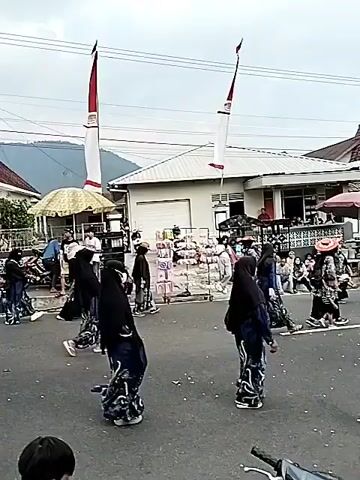 A group of people, many wearing black headscarves and patterned dresses, are walking down a paved street in what appears to be a procession or parade. A large red, blue, and orange flag is prominently displayed in the foreground, attached to a bamboo pole. In the background, there are buildings, stalls, and other people observing the event. Some individuals are holding small flags. The overall atmosphere seems festive, with a crowd gathered to watch.