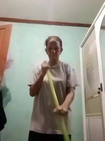 A person is standing in a room and sweeping with a broom. The person is wearing a light-colored t-shirt and dark pants. The room has a light blue wall and a white door on the right side of the frame. There is also a wooden door on the left side of the frame. The lighting is dim.