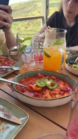 The video shows a table setting with food and drinks. In the foreground, a dish of food that appears to be eggs scrambled with tomato sauce and topped with sliced green peppers and red peppers is presented in a ceramic bowl. A spoon rests in the bowl. To the left, a tall glass with ice and mint leaves, and some pink liquid, is partially visible. To the right, a pitcher filled with orange juice and ice, garnished with mint leaves, sits on the table. In the background, two people are seated at the table. One person on the left is partially visible, wearing a light-colored shirt and holding a phone. The person on the right is more visible, wearing a dark t-shirt, and looking down. The background suggests an outdoor or semi-outdoor setting with greenery visible through a window or opening. The table surface is made of wood.