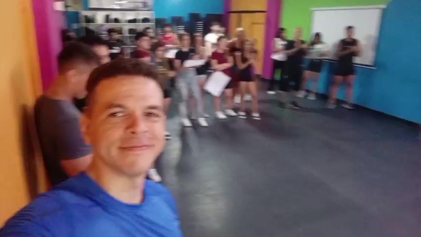 A man takes a selfie in a dance studio, capturing a group of about twenty people behind him. The man, wearing a blue athletic shirt, smiles at the camera. Behind him, the group stands or leans against a wall, facing forward. Some hold certificates. The studio has colorful walls and a black floor. A projector screen hangs on one wall, and shelves can be seen in the background. The overall lighting is bright and even. The sounds of music are in the background.