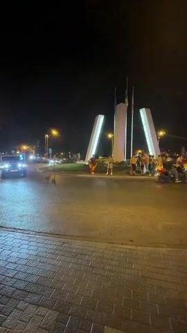 A night scene captures a street with a roundabout. A group of people stand on the roundabout, some holding flags. Several cars, including a white Jeep, a silver sedan, and a white station wagon, are driving through the roundabout. Streetlights illuminate the scene.