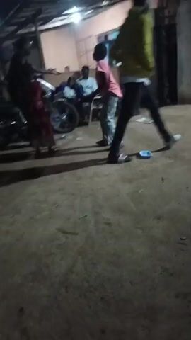 The video shows a person standing outdoors at night. The person is wearing a light-colored long-sleeved shirt, dark pants, and sandals. The person in the foreground appears to be swaying or moving erratically. In the background, several people are sitting on chairs. There are also vehicles and buildings visible in the background, illuminated by streetlights. The lighting is poor, and the overall scene appears blurry due to movement. Another person in a dark t-shirt stands next to the first person, partially blocking the view.