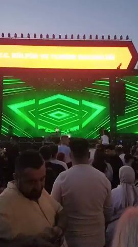 The video shows a concert crowd standing in front of a stage. The stage has a large screen displaying green geometric shapes and a spotlight. A sign above the screen reads "T.C. KÜLTÜR VE TURIZM BAKANLIĞI" which translates to "T.R. MINISTRY OF CULTURE AND TOURISM" in English. The sky is visible above the stage, suggesting it is an outdoor event. The people in the crowd are standing, some looking at their phones, others looking at the stage. The overall atmosphere seems to be one of anticipation before or during a concert.