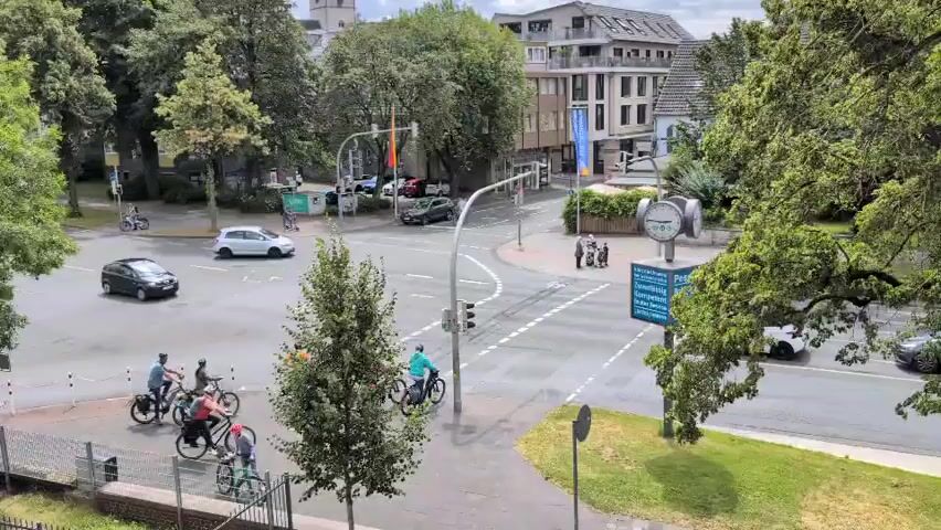 The video shows a wide shot of a street intersection with traffic lights. Several people are riding bicycles in the foreground and middle ground. There are also cars on the road, including a white car, a dark SUV, and other vehicles in the background. The environment appears to be urban, with buildings and trees visible. The weather seems to be overcast, with the sky appearing light gray. A sign with blue lettering is visible on the right side of the intersection. The cyclists are moving in various directions, some stopping at the intersection and others proceeding through. The traffic lights are visible and appear to be operational. A fence and some grass are in the lower left portion of the frame.