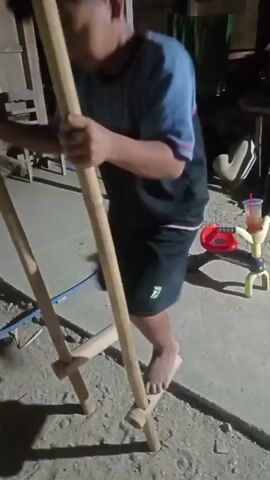 A child wearing a blue and black t-shirt and black shorts is standing on stilts made of wood. The child is holding onto two long wooden poles for balance. The ground appears to be dirt or sand. The lighting is dim, suggesting it is nighttime or indoors. There are some blurry objects in the background, including what looks like a red toy and some wooden structures. The child is in the process of walking or attempting to walk on the stilts.