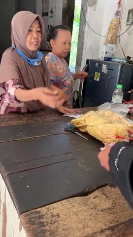 The video opens with a close-up of a wooden counter where a person in a brown hijab is handing money to another person. Behind them, a woman in a colorful shirt is talking and gesturing, with bags of food products hanging in the background. The camera then pans to the left, showing shelves stacked with numerous individually wrapped food items, tied with red string. The environment appears to be a small shop or market stall.