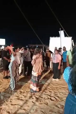 At night, an outdoor scene unfolds on sandy ground, likely a beach or an event with artificial sand. Numerous individuals, a mix of men and women, are gathered under artificial lighting. In the foreground, two women are dancing. The woman on the left wears a white and black patterned outfit and dances barefoot, occasionally raising her arms. The woman facing her on the right wears a striped top and patterned trousers. They briefly hold hands while dancing. Surrounding them, other people stand and observe, some interacting with each other. In the background, large white rectangular structures, possibly set pieces or lighting equipment, are visible. Tall poles and cables extend into the dark sky. A colorful cart stands behind the dancers. A woman in blue jeans and a blue patterned top stands in the extreme right foreground, her back mostly to the camera, observing the scene. Music is audible throughout the clip.