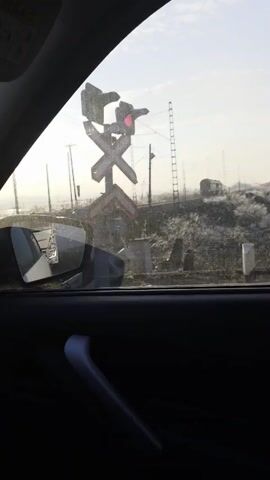From inside a car, a view out the side window reveals a railroad crossing signal flashing red. The sign is silhouetted against a hazy, overcast sky. In the distance behind the sign, a train is visible, parked on the tracks. The surrounding area appears to be a somewhat bleak, rural landscape with utility poles and scattered debris. The car's side mirror and interior door panel are visible in the foreground.
