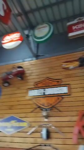 The video shows the interior of a room decorated with vintage signs and memorabilia. There are numerous signs displayed on the wooden walls, including those for brands like Harley-Davidson, Buick Service, Shell Gasoline, Coca-Cola, Marlboro, and Texaco. Above the walls, there are additional round signs hanging from the ceiling, featuring logos of various beverages and products. An American flag is hanging on one of the walls. A yellow crane structure is also visible in the room. In the final shot, a man wearing a black shirt enters the frame, walking towards the back of the room. The overall setting appears to be a themed room, possibly a bar or a recreation area, styled with an Americana aesthetic.