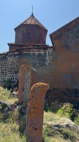 A vertical video clip shows an old stone building with a copper dome and a cross on top. The building is partially covered in orange moss and lichen. In the foreground, two weathered, orange-covered stone markers stand upright. The sky is clear and blue, and there is green grass and some rocks around the base of the building.