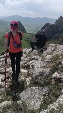 A video shows two people hiking on a rocky mountain trail. The foreground has a woman wearing a pink hat and orange shirt walking towards the camera. Behind her and slightly to the right, a person wearing a dark shirt and backpack is sitting on a rock, looking at the view. The camera then shifts to show that person standing up and walking away towards the right. In the background, there is a valley with green fields, mountains, and a cloudy sky. The terrain is rocky, with patches of grass. Hiking poles are visible on the ground.