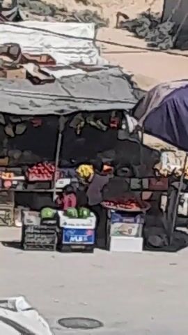 A street vendor stand is visible in the center of the frame. The stand is covered by a tarp and displays various fruits, including oranges, apples, and what appear to be watermelons and strawberries. There are also boxes and crates stacked around the stand. Two men are present. One man, wearing a black shirt with white accents, is standing to the left of the frame, looking towards the stand. The other man, wearing a dark shirt, is walking away from the stand towards the right side of the frame. The background shows a sandy, barren landscape with some structures or tents. The overall scene depicts a typical outdoor market environment.