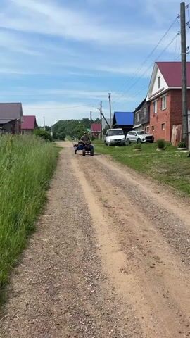 A person is riding a small, blue motorized cart with a trailer on a dirt road in a rural village. The person is wearing a camouflage shirt and a green hat. The cart is moving away from the camera. On the right side of the road, there are houses with red roofs and a wooden fence. On the left side of the road, there is tall grass. The sky is blue with some clouds. The road is unpaved and appears to be made of gravel. There are power lines visible in the background.