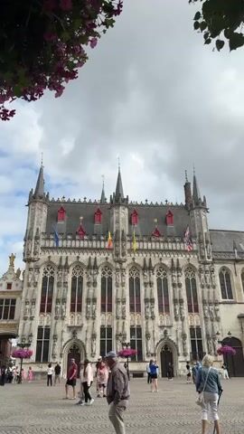 The video shows a wide shot of the exterior of a large, ornate building with multiple spires and arched windows. Flags are displayed on the building. The building is situated in a public square paved with cobblestones. Several people are walking around in the square, and some are standing in small groups. The sky is overcast with clouds. In the foreground, a man is walking towards the camera, and to his right, a woman is walking away from the camera. Pink flowers in hanging baskets are visible on the left side of the frame, and green foliage is visible on the right side. The camera is pointed upwards towards the building.