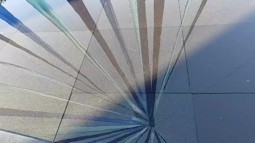The video shows a reflection of a canopy of colorful fabric strips in a water feature in front of a Nike store. The canopy is made up of blue, teal, and dark gray fabric strips, which create a striking visual effect as they are reflected in the water. The Nike store is visible in the background, with its distinctive logo and large glass windows. Several people are seen walking in and out of the store. The water feature appears to be a shallow pool or a decorative pond, with a tiled bottom. The overall scene is one of modern architecture and urban design, with a focus on light, color, and reflection.