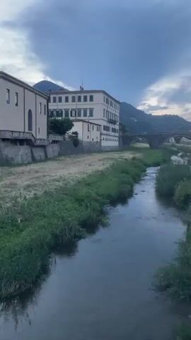 The video shows a narrow river flowing through a town at dusk. On the left side of the river is a multi-story building with a light-colored facade and green shutters. Opposite the building, on the right side of the river, is a grassy embankment with a few bushes and trees. Further in the background, a stone bridge with multiple arches spans the river. Beyond the bridge, there are more buildings and hills covered with trees. The sky is cloudy, with streaks of pink and orange suggesting the sun has recently set. The water in the river is dark and reflects the sky. The overall mood is peaceful and serene.