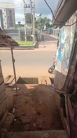 The video shows a street view from a narrow opening. In the foreground, a dirt path leads down to a street. There are wooden planks and a drain visible on the path. On the right side of the opening, there is a wall covered with colorful posters and some buckets. The street itself is paved and appears to be relatively empty. Across the street, there is a modern-looking building with multiple floors and large windows. There are also some trees and streetlights visible in the background. A motorcycle is parked on the side of the road. The sky is cloudy.