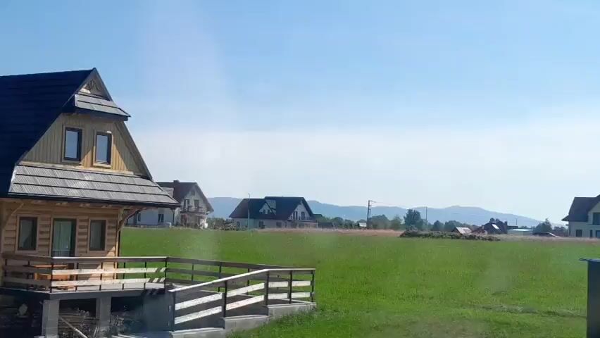 A drone shot shows a wooden house with a dark roof in a grassy field under a clear blue sky. The house has a balcony with a wooden railing and several windows. In the background, more houses and trees can be seen, along with rolling hills. The shot pans slightly to the right, revealing more of the surrounding landscape.