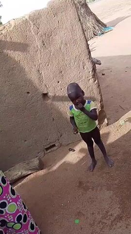 A young, barefoot child stands outside, next to a section of a mud-brick wall. The child wears a green t-shirt and dark shorts. The child’s fingers are near their mouth. A person wearing a pink patterned fabric is partially visible in the lower left corner. The ground is dirt. Behind the wall, a thatched structure is visible.