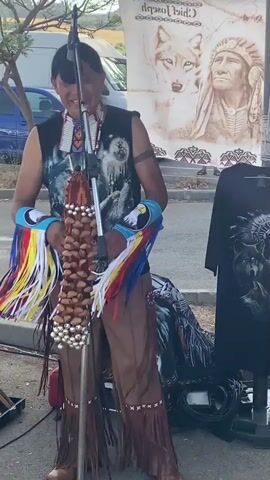 A man dressed in Native American attire, including a headdress, beaded necklace, and fringe, stands in front of a microphone. He holds a musical instrument that appears to be made of gourds and beads. Various pan flutes are displayed on a red cloth in the foreground, and a black t-shirt with a wolf and Native American imagery is on a mannequin in the background. The scene appears to be outdoors in a parking lot, possibly at a market or festival, with cars and trees visible. The man is smiling and seems to be interacting with the microphone.