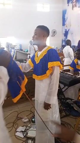 A young man in a white robe and a blue and yellow patterned sleeveless top stands in the center of a room, swaying and moving his hands as if singing or leading worship. He has a faded haircut and a slight goatee. In the background, to the right, another man in a similar blue and yellow top over a white robe is seated and playing an electronic keyboard. An electric guitar rests on the floor nearby. The floor is cluttered with numerous black electrical wires. On the back wall, a large light blue banner displays a white image of Jesus with outstretched arms, along with partially visible white text including "REDEMPTION" and "OWORD ES". Other individuals in white robes are scattered in the background. The room has white walls and a light-colored floor, suggesting a church or worship hall. A white plastic chair is partially visible in the foreground to the right.