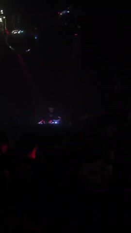 This media file captures a vibrant scene inside a nightclub. The primary subjects are a crowd of people, a DJ at a console, and a prominent neon sign. The setting is a dimly lit venue, bathed in a strong red light emanating from the stage and amplified by disco balls hanging from the ceiling. The atmosphere appears energetic and lively, with the crowd seemingly engaged in dancing and enjoying the music, as suggested by their postures and expressions. The DJ is actively working, indicated by their position behind the equipment, and the pulsating red lights create a sense of excitement and immersion.

The visible text on the neon sign reads "LA JUNTA UNIVERSITARIA," suggesting that this event is hosted or organized by a university group or society. The overall activity is a music event or party, typical of a nightclub environment. The time of day is implied to be night, consistent with the typical operating hours of such venues and the artificial lighting used. There are no indications of weather conditions as the scene is entirely indoors. The location context of Iquique, Chile, adds a specific geographical detail to the setting. Emotions conveyed through the visuals are primarily exhilaration and enjoyment, driven by the music, lighting, and social gathering.