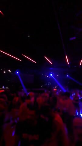 The video shows a crowded nightclub with a dark ceiling adorned with red neon lights. The room is illuminated by blue laser beams and red and purple club lights, creating a vibrant and energetic atmosphere. A large group of people are dancing, their bodies silhouetted against the colorful lights. The camera pans across the scene, capturing the movement and excitement of the partygoers. Some individuals are visible clearly, while others are blurred by the motion and low light conditions. The overall impression is of a lively and immersive nightlife experience.