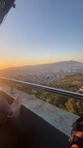 The video captures a panoramic view of the city of Tétouan, Morocco, at sunset from a high vantage point, likely a balcony or terrace. The initial shot shows a person's feet, clad in black sandals, resting on a metal railing in the foreground. The camera then pans to the right, revealing an expansive urban landscape stretching across a valley, bordered by mountains on the horizon. The sky is clear, displaying the warm, soft colors of dusk. Below the viewpoint, green fields, scattered houses, and cultivated areas are visible, transitioning into the denser city environment. A vertical metal pole momentarily obstructs part of the view during the pan. The scene suggests a tranquil moment, enjoying the vast and picturesque landscape as the day ends.