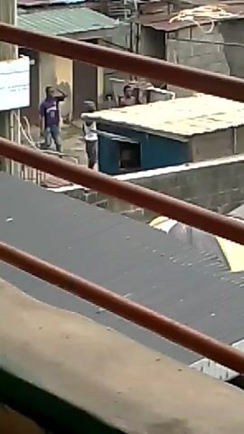 From an elevated perspective through orange railings, two men are visible in a densely populated area with buildings made of various materials, including corrugated metal. One man wears a purple shirt and jeans, while the other has on a white tank top and shorts. The man in the tank top appears to be gesturing or pointing. The buildings appear to be in a state of disrepair.