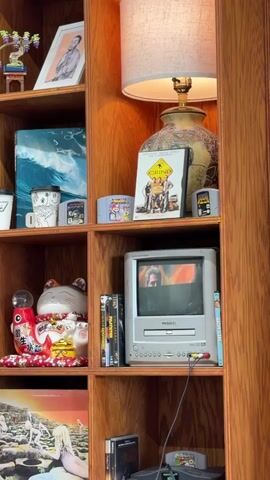 The video shows a close-up view of a wooden bookshelf filled with various items. On the top shelf, there is a small decorative sculpture, a framed picture, and a lamp with a patterned lampshade. The second shelf from the top contains a framed picture, a mug, and several video game cartridges. The third shelf holds a ceramic lucky cat statue, a stack of DVDs, and a small CRT television displaying an image. The bottom shelf features a vinyl record and a video game console. The camera pans slightly to the right, focusing on the items on the shelves. The lighting appears to be coming from the lamp on the shelf and ambient room lighting. The overall environment suggests a home or personal collection display.