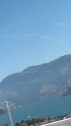 A paraglider is seen soaring through a clear blue sky above a vast lake and a mountainous landscape. The paraglider, a small white and blue canopy with a dark figure suspended beneath it, appears to be in mid-flight, moving across the frame from left to right. Below the paraglider, the expansive, blue-green lake stretches out, dotted with a few white boats. The distant shores of the lake are lined with trees and houses, climbing up the steep, green slopes of the mountains. The mountains themselves are rugged and imposing, their peaks partially obscured by a hazy mist. The sky is clear except for a few wispy white contrails from an unseen aircraft, forming an 'X' shape. The overall impression is one of a serene and adventurous day, with the paraglider the focal point of human activity against a majestic natural backdrop.