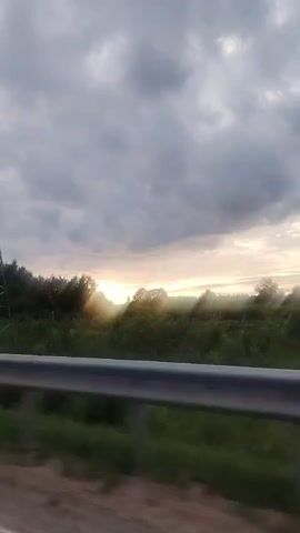 This is a video recording taken from a moving vehicle in Saurieši, Latvia. The footage captures a landscape scene with a prominent electrical tower in the foreground on the left side of the frame. Beyond the tower, a line of trees and vegetation borders a field. The sky is filled with dramatic, overcast clouds, but rays of sunlight are breaking through, suggesting either sunrise or sunset. The presence of the electrical tower and the general natural setting indicate a rural or semi-rural environment. The motion blur suggests the video was filmed while traveling, likely on a road. There is no visible text in the video. The overall mood is serene and somewhat dramatic due to the sky's appearance.
