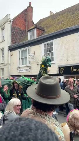 The video captures a vibrant parade taking place on a street lined with buildings, with a large green dragon puppet as a central figure. The dragon is decorated with foliage and sunflowers, and appears to be operated by multiple people. Spectators line the street, some dressed in green attire, with others donning flower crowns. A man in a green hat is captured in the foreground, with a woman in gray standing adjacent to him. Individuals are seen taking photos and videos of the parade with their mobile phones. Building signage is visible, including "Roberts Curios" and "Through the Looking Glass." The overall atmosphere is festive and lively.