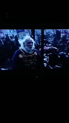 The video shows a scene from a historical film, possibly a battle. In the foreground, an elderly man with white hair and a beard, wearing a blue cloak and ornate armor, is visible. He appears to be a leader or a figure of authority. Behind him, a large group of people, likely soldiers, are engaged in combat. Red flags and banners are prominent in the background. The lighting is dramatic, with blue and purple hues dominating the scene, suggesting a nighttime or twilight setting. The overall atmosphere is chaotic and intense.