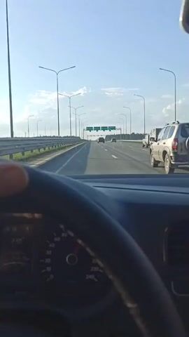The video shows a view from inside a car, driving on a highway. The driver's hands are visible on the steering wheel. The dashboard is in view and shows the car's speed. There are multiple signs overhead indicating directions to Perm. Other cars are visible on the highway ahead. The sky is blue with some clouds and the sun is shining. The environment appears to be rural.