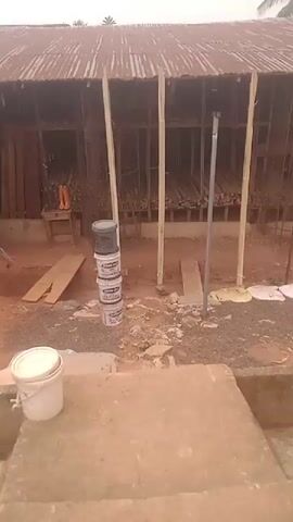 The video shows an outdoor construction site. The ground is covered in a mix of gravel, dirt, and red clay. In the foreground, there is a large concrete block or step, with a white bucket on top of it. Further back, there are four stacked paint buckets. Behind them is a wooden structure with a corrugated metal roof, which seems to be used for storing long, thin pieces of wood or metal, possibly rebar. Inside the structure, there are multiple shelves. The structure has vertical wooden supports and appears unfinished or under construction. There is also a metal ladder leaning against the side of the structure. The video pans slightly to the right, showing more of the construction site. In the background, there is a glimpse of a palm tree.