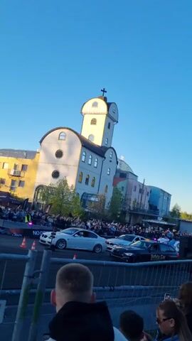 The video captures an outdoor car event on a clear, sunny late afternoon or early evening. A large crowd of people, mostly adults, stands behind metal barriers, attentively observing the scene. Many individuals in the foreground are holding up their phones to record.

In the mid-ground, several cars are visible on what appears to be a track or staging area. A white BMW sedan is prominent on the left, while a black car clearly marked "Safety Car" is positioned towards the center. Other vehicles are lined up further down the area.

Dominating the background is a distinctive multi-story building with a light-colored facade and a tall, cylindrical tower topped with a cross. This unique architecture suggests a church or chapel, fitting for the Geiselwind, Germany location. Lush green trees and foliage are visible beyond the building and along the perimeter of the event space. The sky is a bright, cloudless blue, indicating pleasant weather conditions. The overall atmosphere suggests an engaging public event centered around automotive activities.
