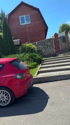 The video begins with a side view of a red Mazda 3 parked on a street. The camera pans to the right to show the front of a white SUV parked next to it. The camera then moves forward to show a group of people walking towards a building with large windows and a modern design. The video pans again to the right to show a black car parked next to a large SUV, with a river and trees in the background. The video ends with a close-up of the front grille of a white Mercedes-Benz. The scene is set on a sunny day with a blue sky and some clouds.