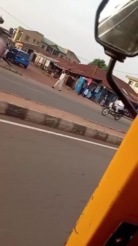 The video, shot from inside a yellow vehicle, possibly a tricycle, shows a view of a road in what appears to be a town or small city.  There are various types of vehicles on the road, including cars and motorcycles.  The streets are lined with small businesses, some with open fronts. Pedestrians are visible, including a man in a white robe. The sky is overcast. The camera vehicle moves along the road, capturing passing traffic and the streetscape. A few utility poles with streetlights are also visible in the distance.